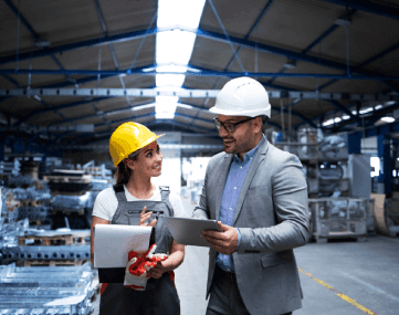 Male and female engineers in hard hats collaborating and reviewing documents on a tablet inside a manufacturing facility